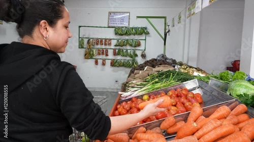 A woman is shopping in a fruit and vegetable store. He selects a carrot and places it in his market cart
