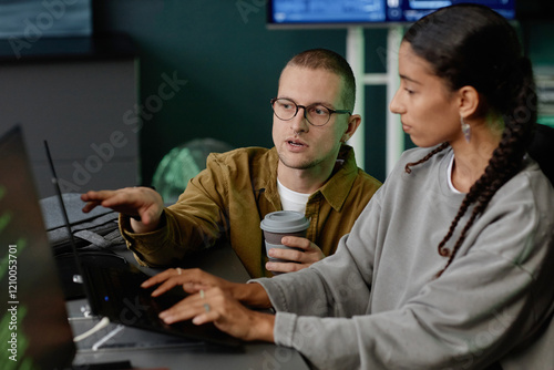 Portrait shot of male IT programmer with glasses pointing at laptop screen while consulting female colleague during discussion at work meeting in office
