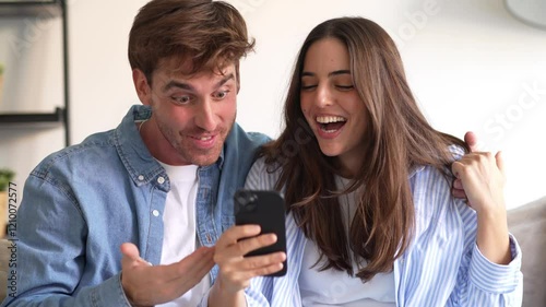 Excited millennial husband and wife relax on couch in kitchen feel euphoric win lottery online on cellphone, overjoyed young couple triumph celebrate read good unexpected news on smartphone gadget
