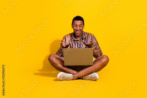 Happy young man using a laptop on a vibrant yellow background, showing a surprised expression and wearing a trendy casual outfit photo