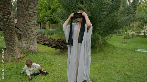 Mother playfully ties blindfold over her eyes and reaches out to catch her son in lush garden of family estate. Boy laughs and dodges her arms, creating joyful moment filled with fun and bonding