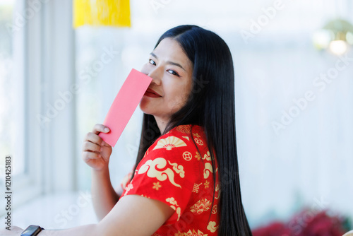 Portrait of happy asian woman relaxing wearing red cheongsam dress holding red envelopes at home in the living room indoor decoration for Chinese new year festival celebrate culture of china holiday