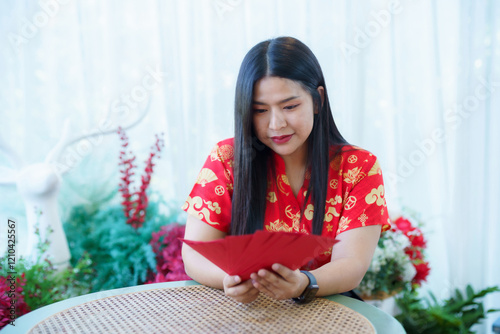 Portrait of happy asian woman relaxing wearing red cheongsam dress holding red envelopes at home in the living room indoor decoration for Chinese new year festival celebrate culture of china holiday