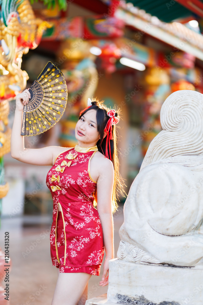 Portrait beautiful cute smiles Asian young woman wearing red traditional Chinese cheongsam decoration and holding a Chinese Fanning for Chinese New Year Festival at Chinese shrine in Thailand