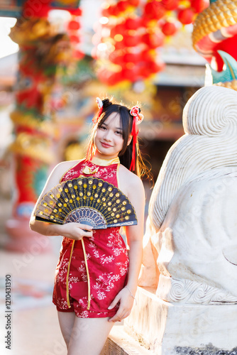 Portrait beautiful cute smiles Asian young woman wearing red traditional Chinese cheongsam decoration and holding a Chinese Fanning for Chinese New Year Festival at Chinese shrine in Thailand