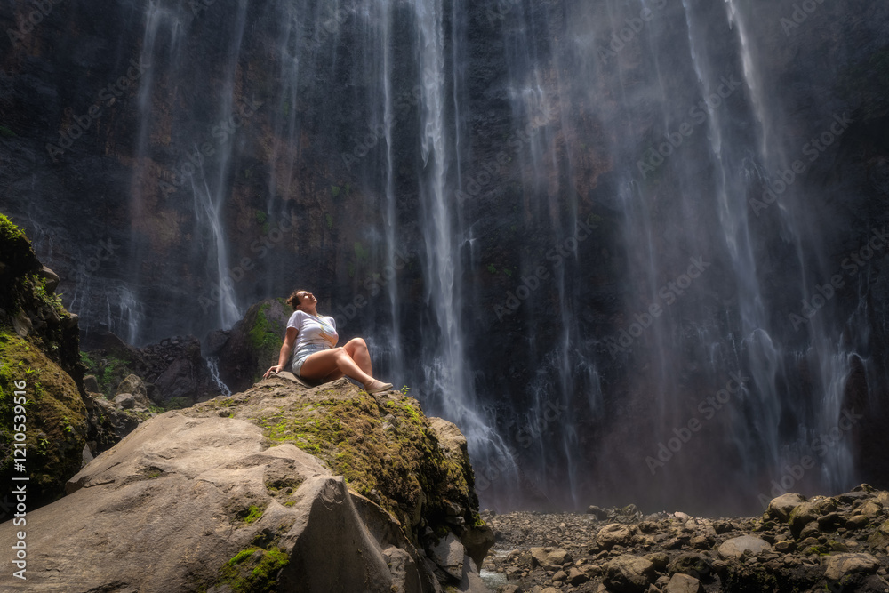 A solitary figure takes a moment to relax by a breathtaking Tumpak Sewu waterfall, completely surrounded by the mesmerizing beauty of nature and soft mist in a peaceful setting, East Java, Indonesia