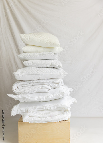 A neatly stacked pile of white pillows, quilts, and bedding arranged against a soft, neutral fabric backdrop. The monochromatic composition highlights textures and layers.