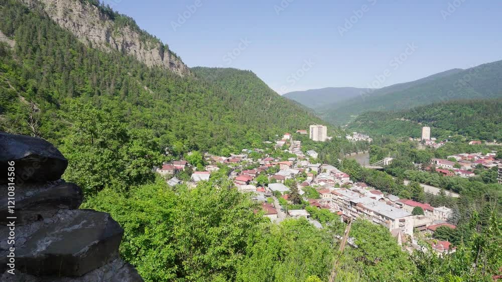 Panoramic view of borjomi city nestled in valley between green mountains under clear blue sky, revealing urban landscape from elevated rocky vantage point