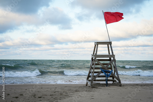 A lifeguard tower stands on a sandy beach as a red flag flutters in the breeze under overcast skies, highlighting safety and vigilance at the waterfront.
