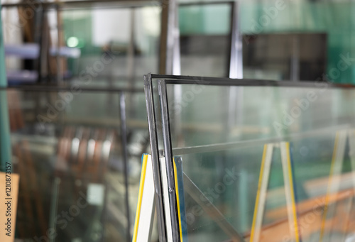 Close-up of insulated glass units stacked on support stand in workshop, with various glass panels and industrial equipment in blurred background