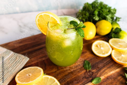 A refreshing green lemonade served in a glass with ice, garnished with mint and lemon slices, placed on a wooden board with fresh lemons and herbs in the background.
