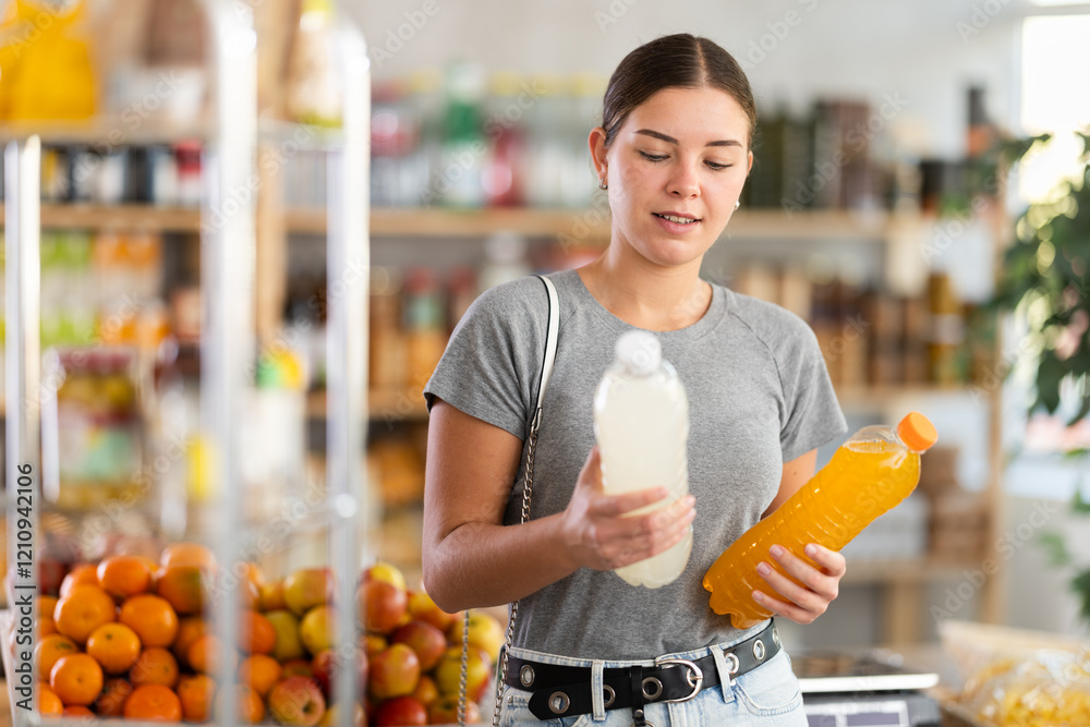 Fototapeta premium While shopping in grocery store, girl buyer choosing drink. Client look package of fruit drink, inspect product, reflect before buying. Customer examines contents of package, chooses beverage