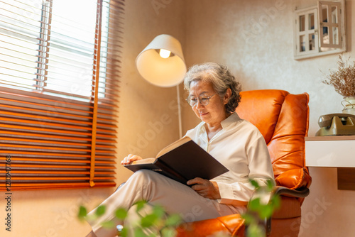 Wellness, mental healthcare in elderly people at home. Happy Asian senior mature woman relaxing on sofa in living room and reading a book. Retired woman enjoy leisure activity hobbies at nursing home.