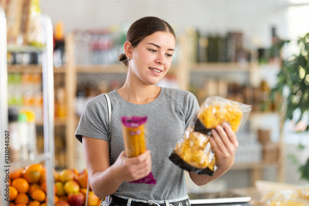 Youth girl choose and buy pasta in store. Market visitor consider and make choice of assortment, buy noodles. Client hesitates before buying, choose garnish product, reads package labels Youth girl choose and buy pasta in store. Market visitor consider and make choice of assortment, buy noodles. Client hesitates before buying, choose garnish product, reads package labels