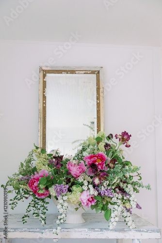 Floral arrangement in white textured compote with pink peonies, irises spirea, tulips, hydrangeas, lilacs on a table in front of a mirror