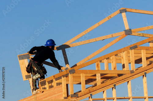 Construction worker in a blue helmet assembling wooden roof trusses on a house frame under a clear sky. Represents home construction, framing, and structural building processes.