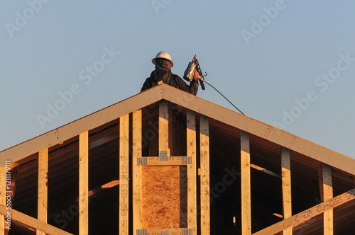 A construction worker in protective gear stands atop a wooden house frame roof under construction, holding a nail gun connected by a hose, illuminated by the warm light of sunrise