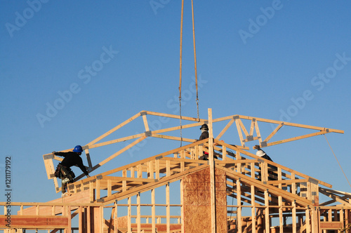 Construction workers install prefabricated wooden roof trusses on a framed house under clear skies, showcasing residential building techniques using cranes and skilled labor in structural assembly