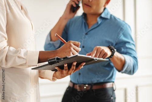 Two professionals discussing businesss and work. The male, of Asian ethnicity, busy on the phone. The female professional is taking notes. Female leader signing a business contract. Working together. photo