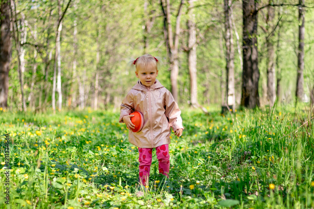 A little girl of 2-3 years old stays in the forest and enjoys nature. Background of a green forest. The girl holds tightly in her hand an orange jar in which she collects the flowers she likes.