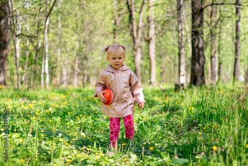 Obraz na płótnie A little girl of 2-3 years old stays in the forest and enjoys nature. Background of a green forest. The girl holds tightly in her hand an orange jar in which she collects the flowers she likes. w salonie