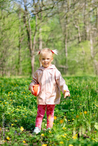 Obraz na płótnie A little girl of 2-3 years old stays in the forest and enjoys nature. Background of a green forest. The girl holds tightly in her hand an orange jar in which she collects the flowers she likes. w salonie