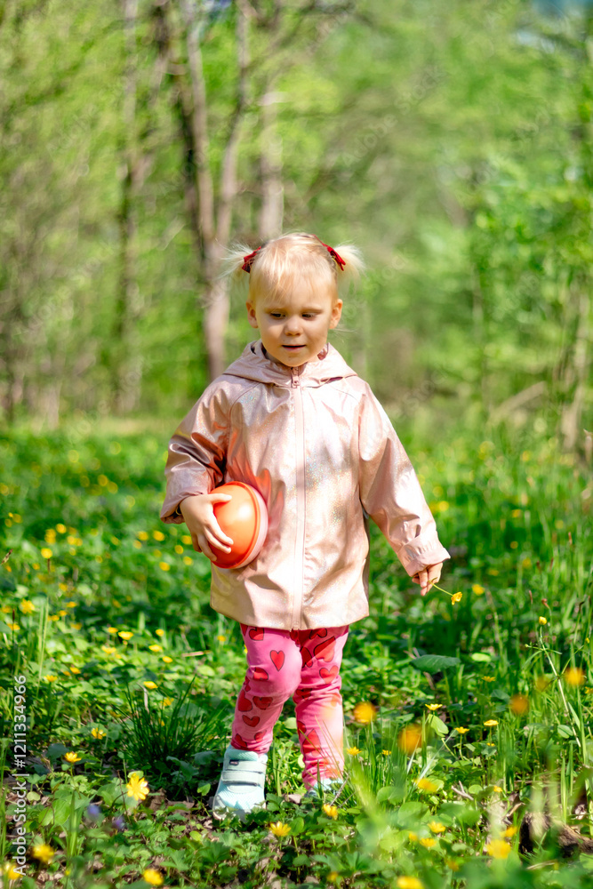 A little girl of 2-3 years old stays in the forest and enjoys nature. Background of a green forest. The girl holds tightly in her hand an orange jar in which she collects the flowers she likes.