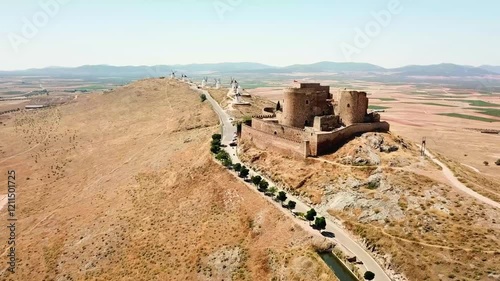 Aerial view of the medieval Consuegra Castle and its iconic windmills standing on a hilltop, overlooking the vast, dry plains of Castilla–La Mancha, Spain, a timeless symbol of Spanish heritage