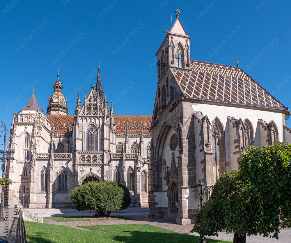 The image shows St. Elisabeth Cathedral in Košice, Slovakia, a stunning Gothic structure with intricate stone detailing, pointed arches, and towering spires. The cathedral stands against a clear blue The image shows St. Elisabeth Cathedral in Košice, Slovakia, a stunning Gothic structure with intricate stone detailing, pointed arches, and towering spires. The cathedral stands against a clear blue