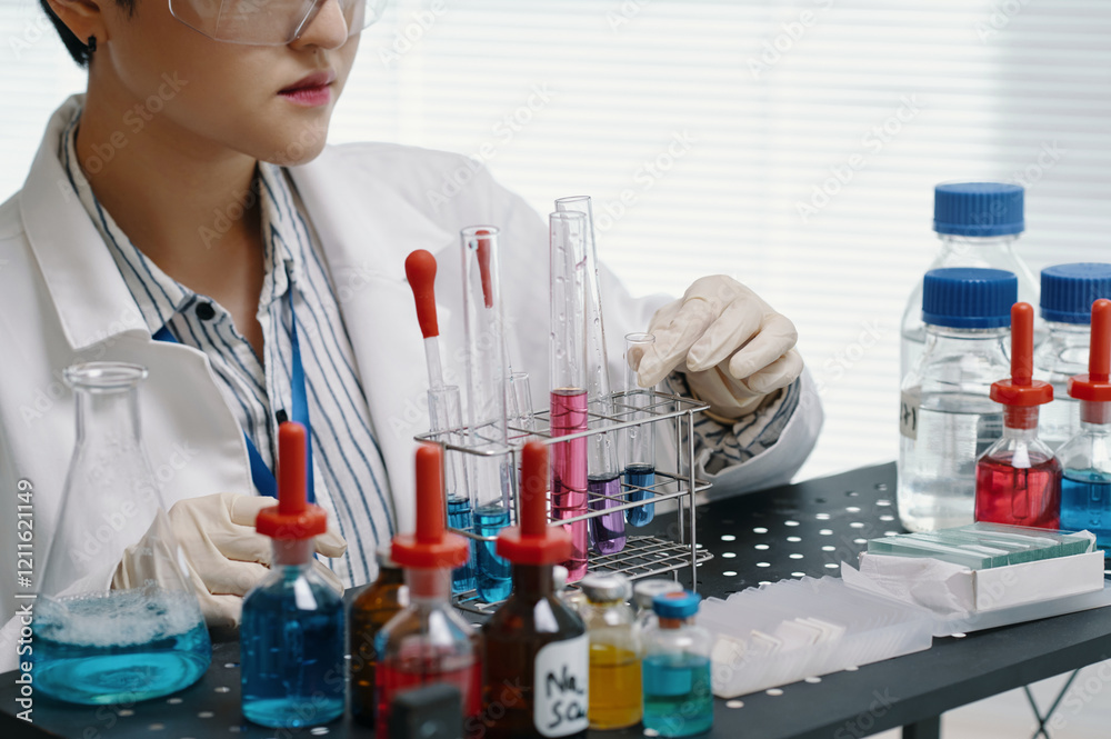 Fototapeta premium Scientist conducting research in lab setting while inspecting chemical solutions and instruments. Multiple bottles and test tubes filled with colorful liquids surround workspace