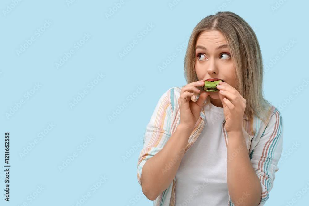Beautiful young woman eating green sweet macaroon on blue background Beautiful young woman eating green sweet macaroon on blue background