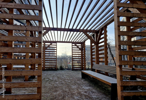 wooden pergola with striped planks on walls and roof. serves as a trellis for climbing plants. sandy beige surface. paths in park with wire benches. foundation rods with screws and threads, winter photo