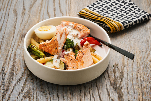 A hearty salmon bowl features freshly cooked salmon, colorful vegetables, and a creamy sauce, served in a white bowl with a black spoon at a cozy dining setting photo