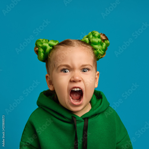 Portrait of emotive child, girl in green hoodie and buns hairstyle shouting, expressing anger and dissatisfaction against blue studio background. Concept of childhood, emotions, lifestyle photo