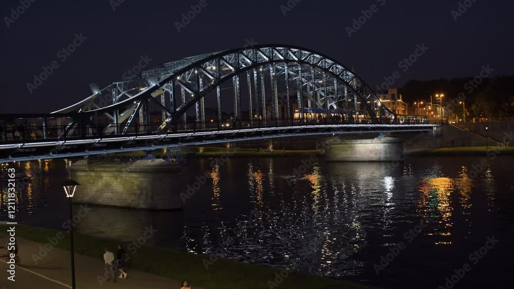 Bridge illuminated at night with reflections in river and people enjoying the riverside stroll Bridge illuminated at night with reflections in river and people enjoying the riverside stroll