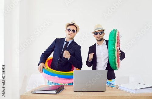 Funny happy company employees standing in office on their workplace with rubber ring and going on holiday trip together. Male coworkers ready for summer vacation. Travel and journey concept.