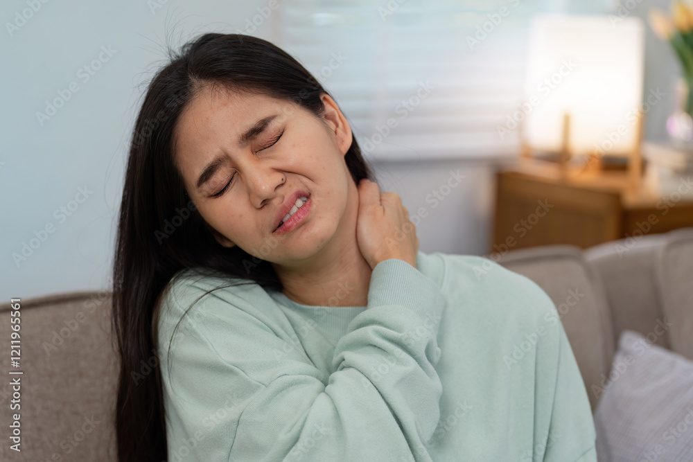 Woman in a mint green sweater grimacing in pain at home, clutching her neck to relieve discomfort and express distress Woman in a mint green sweater grimacing in pain at home, clutching her neck to relieve discomfort and express distress