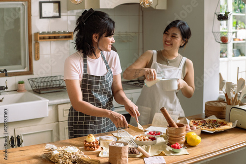 Two Asian women enjoying a baking session in a cozy kitchen. One is cutting waffles while the other pours milk into a bowl. The wooden table displays finished dishes like waffles and pizza
