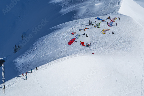 Paragliders prepare for flight from the snowy peaks of Chamonix, seen from Aiguille du Midi. Vibrant parachutes are spread on the ground as adventurers get ready.