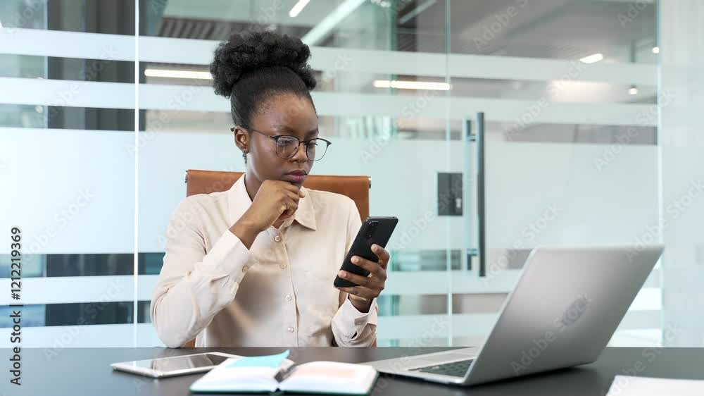 Serious thoughtful african american businesswoman is using mobile phone while sitting in a modern business office. Focused black woman manager works in application, typing a message or chatting online