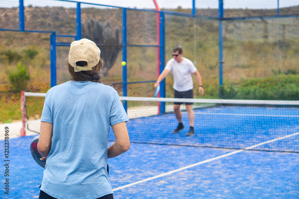 A woman standing with her back to the camera, poised and ready to return the ball on an outdoor padel court,