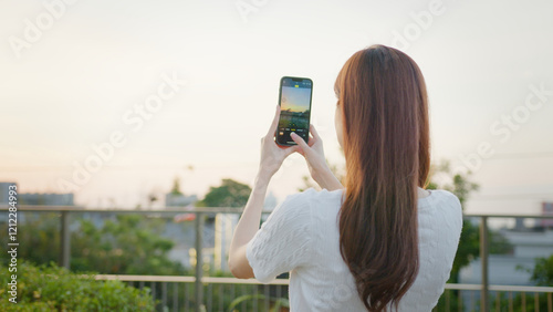 A woman with long hair is taking photo of sunset using her smartphone while standing outdoors on balcony with greenery and cityscape in background. businesswoman digital lifestyle