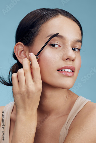 Young woman applying eyebrow gel in a soft, clean setting, showcasing natural beauty and confidence with a pastel blue background