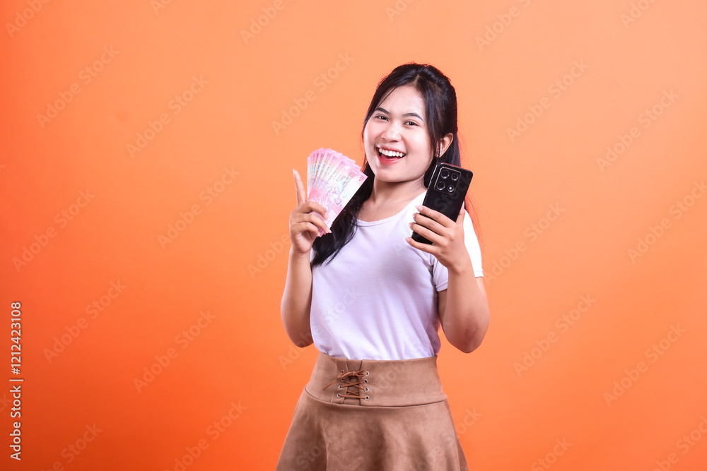 Obraz premium Smiling young woman holding a mobile phone and money against an orange background