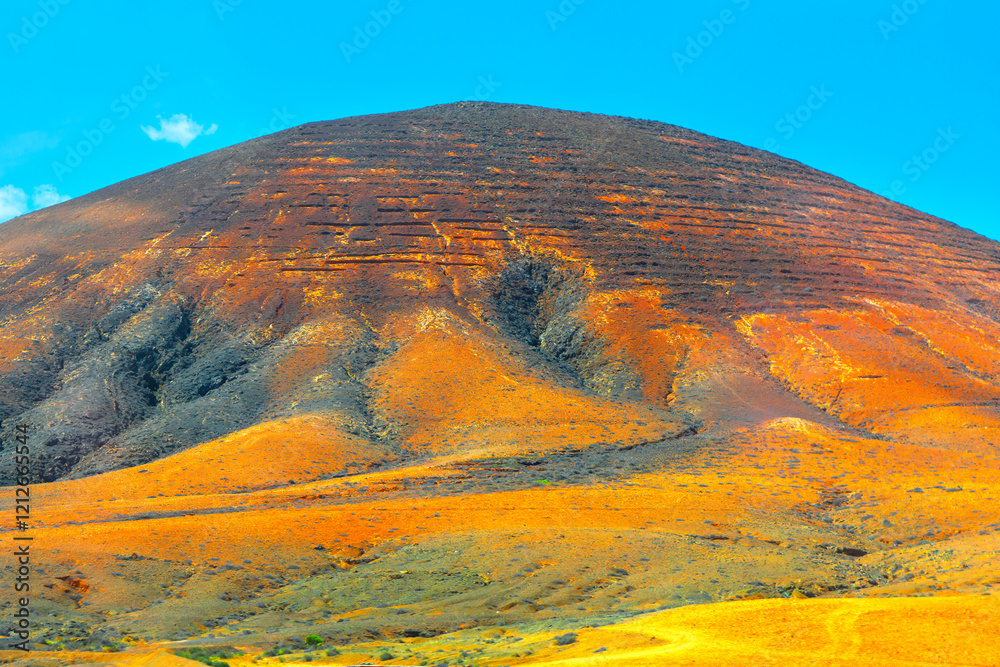 Barren mountain with a distinct reddish hue, set against a bright blue sky. Visible terracing or erosion patterns on the hillside, which create a striking contrast with the vibrant colors of landscape