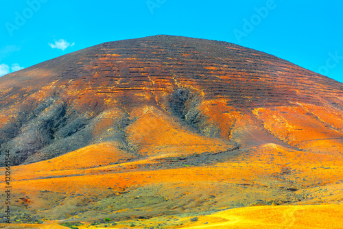 Obraz na płótnie Barren mountain with a distinct reddish hue, set against a bright blue sky. Visible terracing or erosion patterns on the hillside, which create a striking contrast with the vibrant colors of landscape w salonie