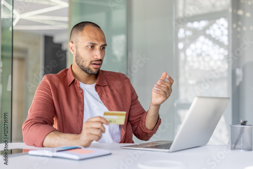 Upset and deceived man at workplace holding bank credit card in hands, received error of funds transfer blocking account, fraudulent actions. Working inside office with laptop.