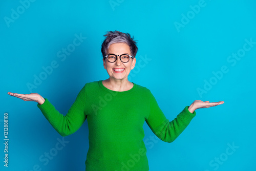 Cheerful elderly woman with grey hair and glasses in green sweater posing with open arms on blue background
