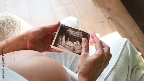 Close up shot side view of unrecognizable pregnant woman admiring sonography picture of her unborn baby sitting at home. Enjoying pregnancy female