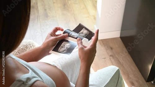 Close up shot side view of unrecognizable pregnant woman admiring sonography picture of her unborn baby sitting at home. Enjoying pregnancy female